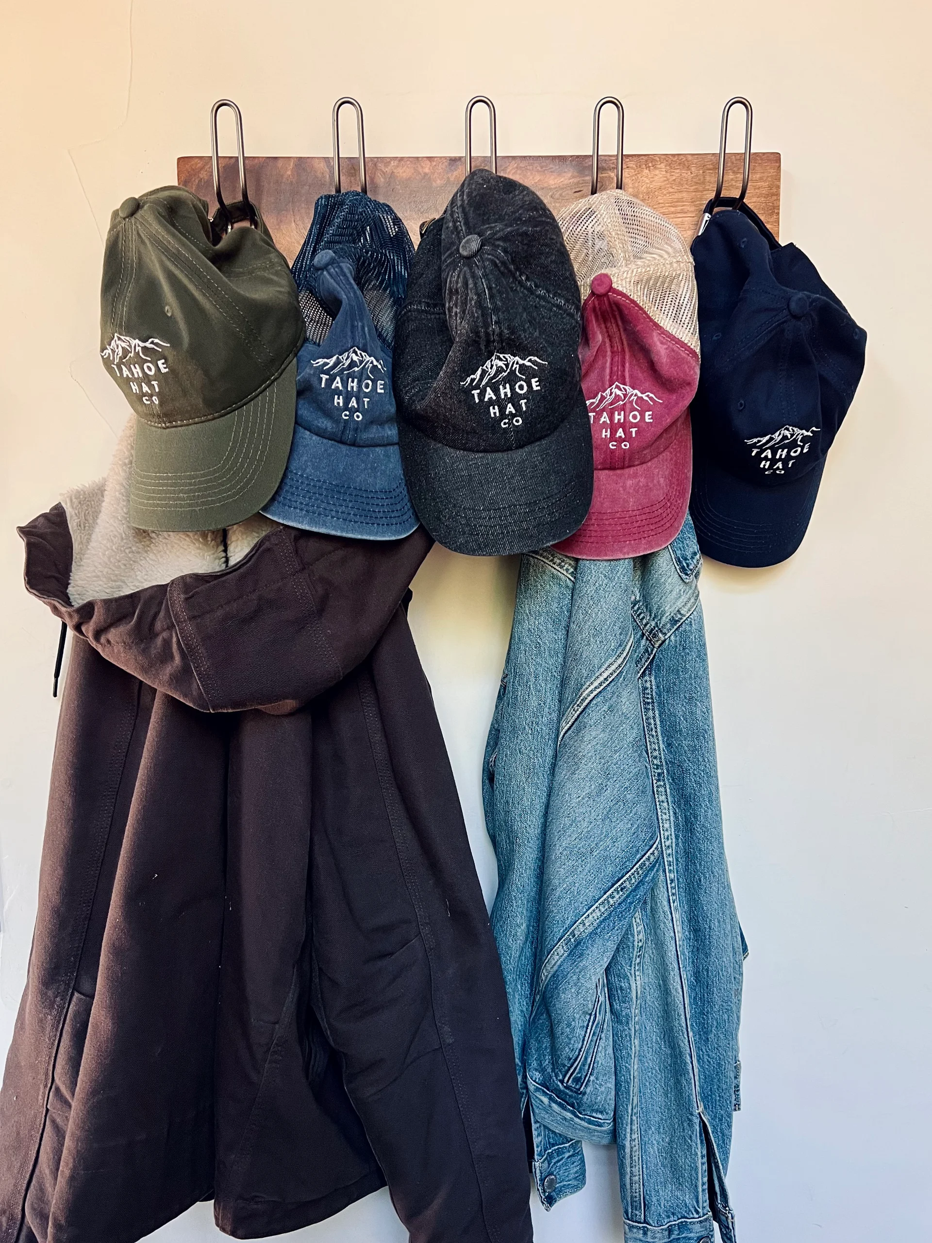 Five Tahoe Hat Co mountain caps hanging on a wooden coat rack above a jacket and denim shirt