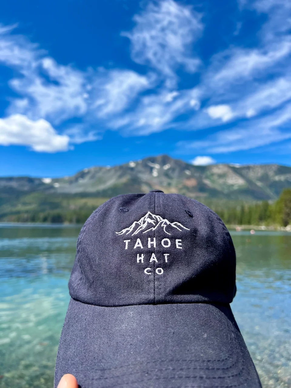 Navy Tahoe Hat Co mountain cap beside clear lake water, with mountains and clouds behind it